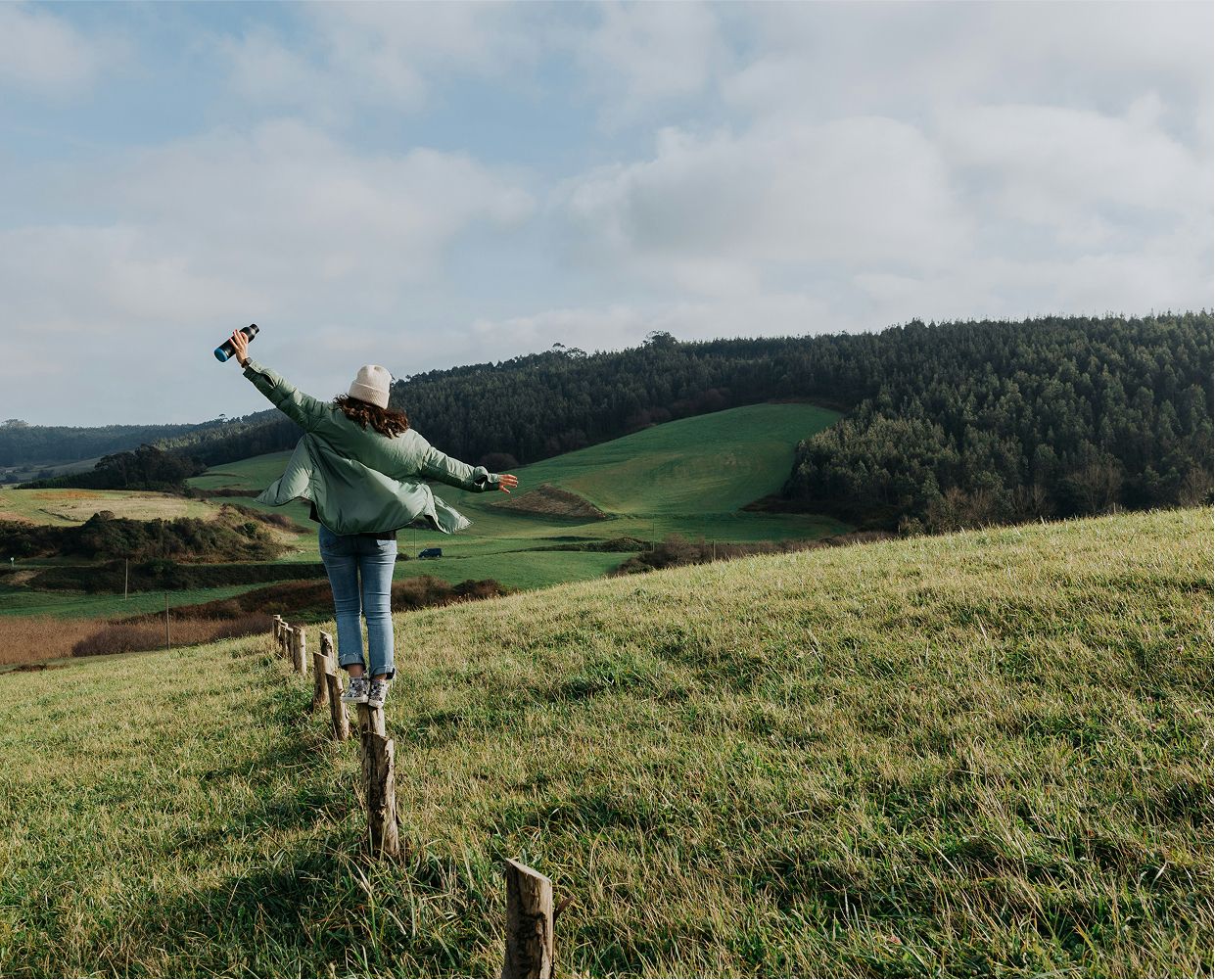 A person stands with arms outstretched on wooden posts in a grassy field, embracing sustainability while overlooking rolling green hills and a forest under a partly cloudy sky.