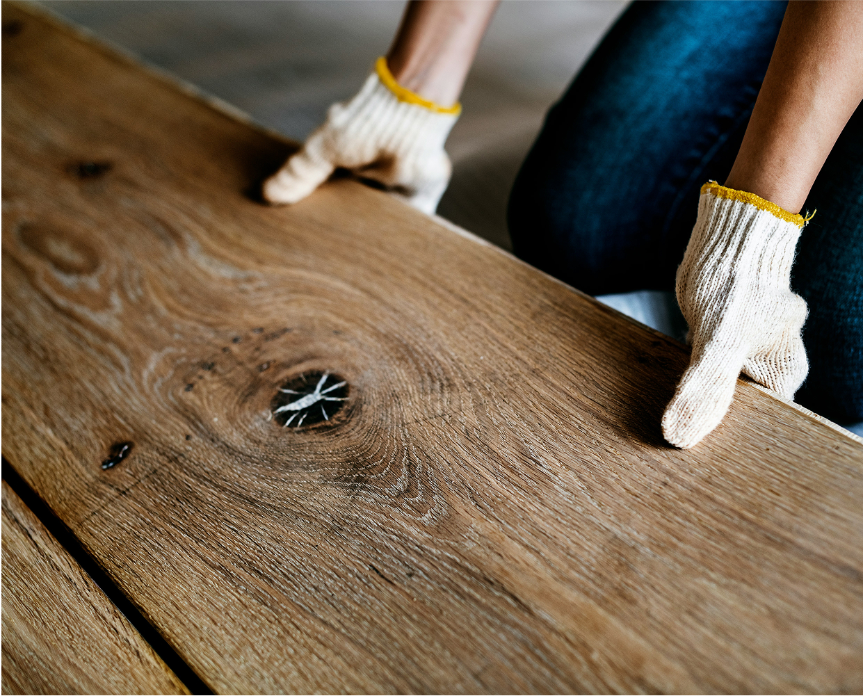 A person wearing white knit gloves is holding a large wooden plank with a visible knot and crack, suggesting sustainable carpentry or furniture making.