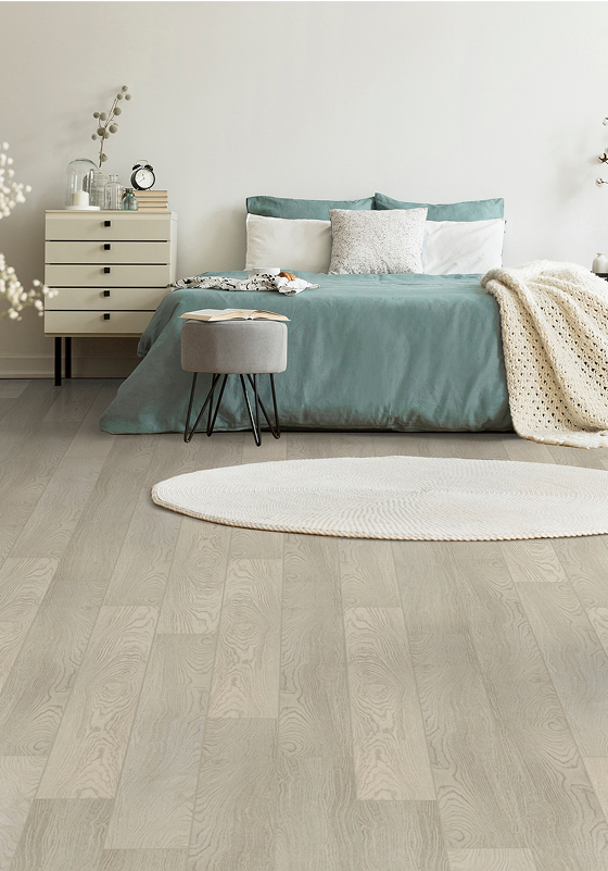 A modern bedroom with a teal bed, white bedding, a cream knit blanket, a round white rug, a grey stool, a light wood floor, a small dresser with decor, and soft, neutral wall colors.