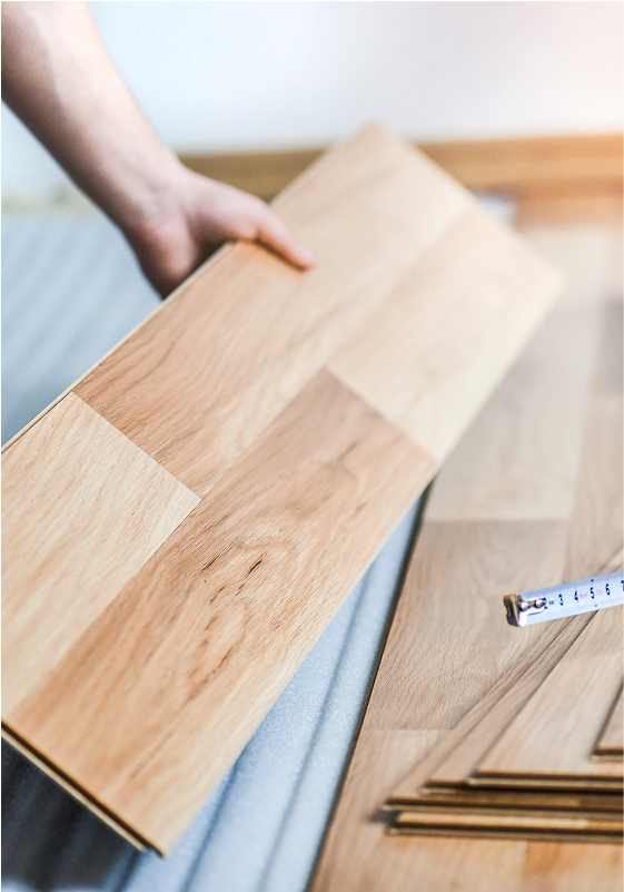 A person holds a light wood laminate plank, preparing to install it on a floor. Other planks and a measuring tape are visible nearby.