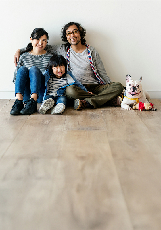 A smiling family of three and a French Bulldog sit on a wooden floor against a white wall, with the parents arms around the child and the dog wearing a colorful outfit.