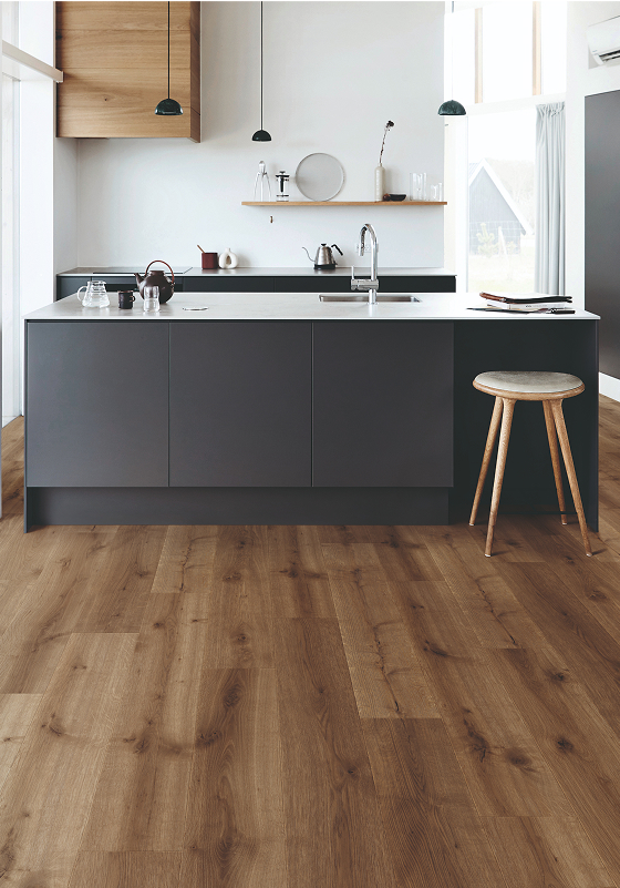 Modern kitchen with a large island featuring dark gray cabinets and a white countertop, a wooden stool, wood-look flooring, and minimalist decor in a bright space with ample natural light.