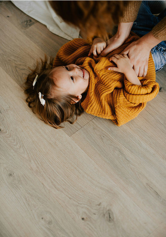 A young girl with brown hair and white hair clips lies on a wooden floor, smiling as an adult gently tickles her. She is wearing a mustard yellow sweater and appears happy and playful.