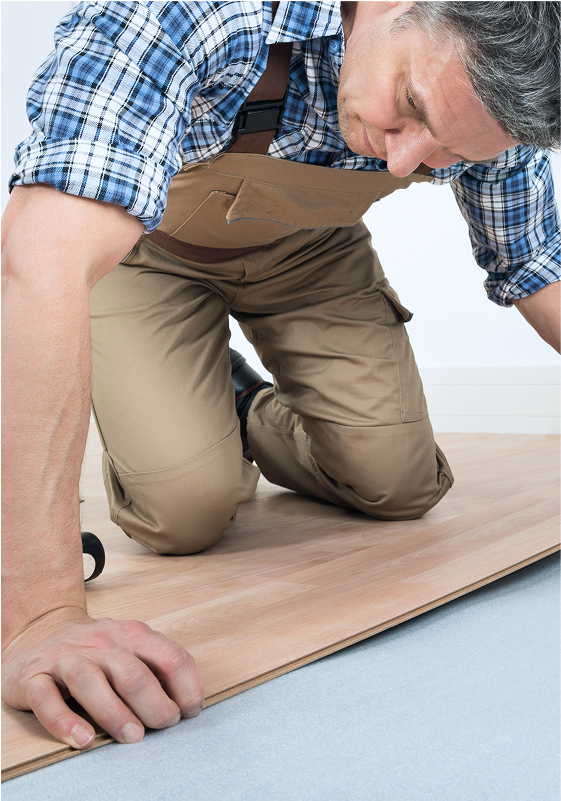 A man in tan work overalls and a blue plaid shirt kneels on the floor, installing a wooden plank as flooring. He is focused on aligning the plank, with tools visible nearby.