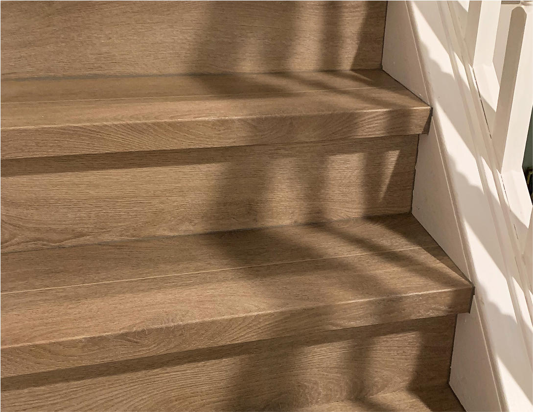 Close-up of three wooden stair steps with a natural finish, enhanced by Smart Floors Accessories. Shadows from a railing create diagonal lines across the steps, and a white wall borders the staircase on the right side.