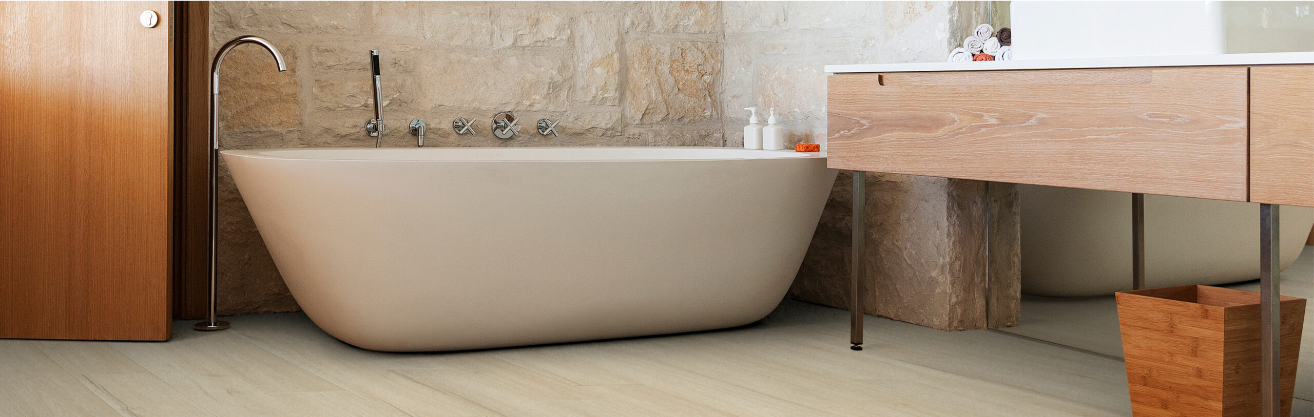 A modern bathroom designed by expert builders features a freestanding oval bathtub, stone wall, wood vanity with a white countertop, and minimalist decor. Bottles and toiletries are placed near the tub.