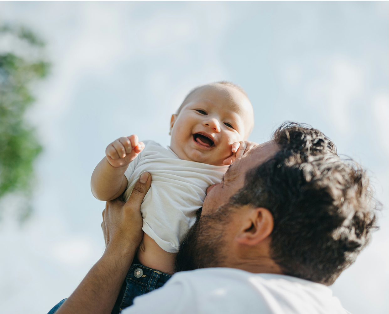 A man lifts a smiling baby into the air outdoors on a bright day. The baby wears a white shirt and looks happy. The sky is clear in the background with some greenery visible.