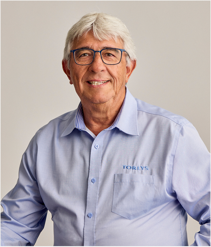 An older man with white hair and glasses smiles at the camera. He is wearing a light blue button-up shirt with TORLYS embroidered on the chest pocket, representing TORLYS flooring. The background is plain and neutral.