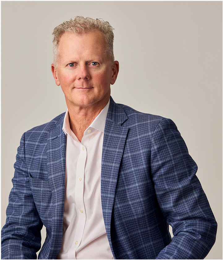 A middle-aged man with short, light hair wearing a blue plaid suit jacket and white shirt sits against a plain, light background, looking directly at the camera with a neutral expression—perfect for showcasing TORLYS flooring style.