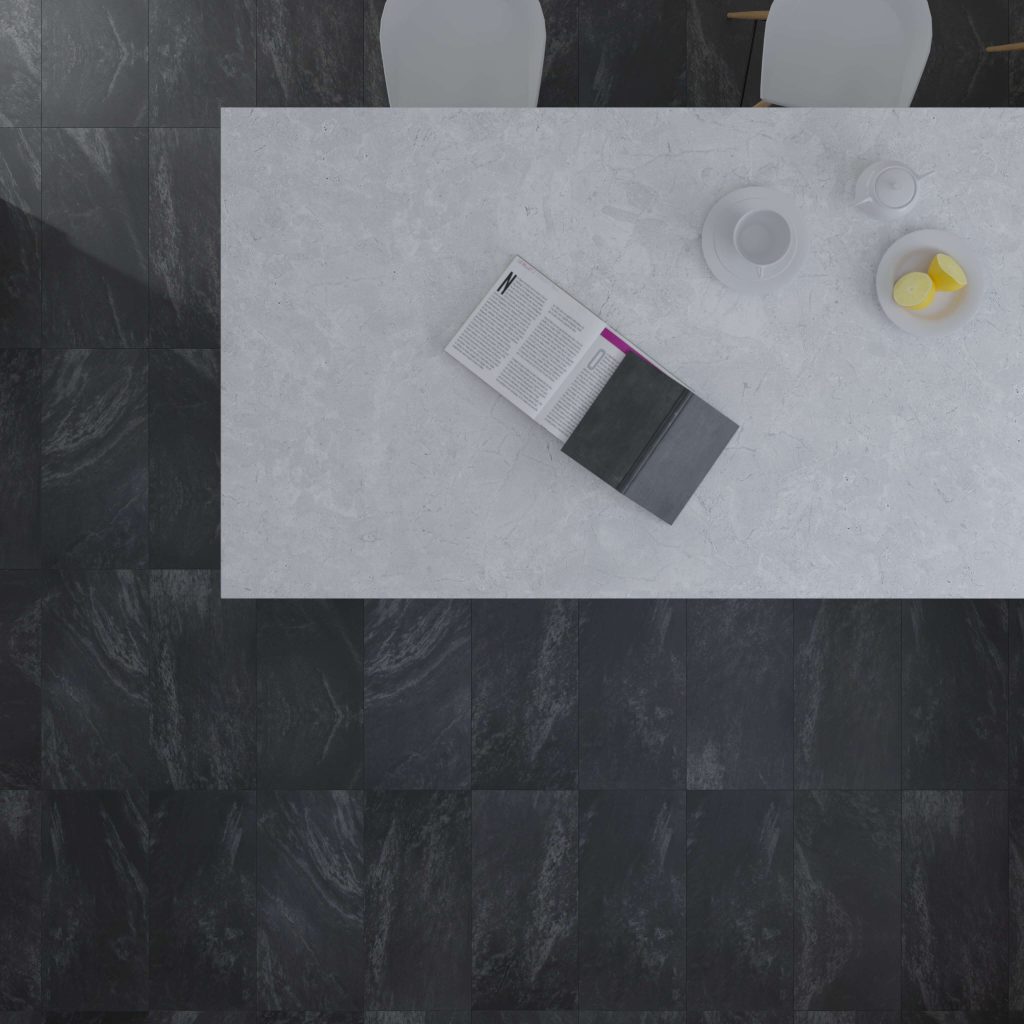 A top-down view of an Anthracite table with an open book, a cup, a teapot, and a bowl of sliced lemon, surrounded by two white chairs on a dark tiled floor.