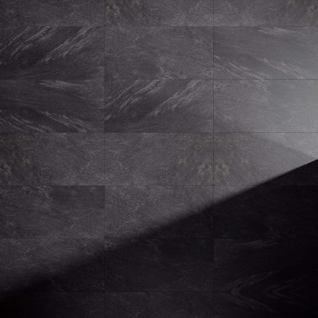 Anthracite tiles with a matte finish are lit by a diagonal beam from the lower right, highlighting their subtle natural textures and patterns, and creating a striking contrast between illuminated and shadowed areas.
