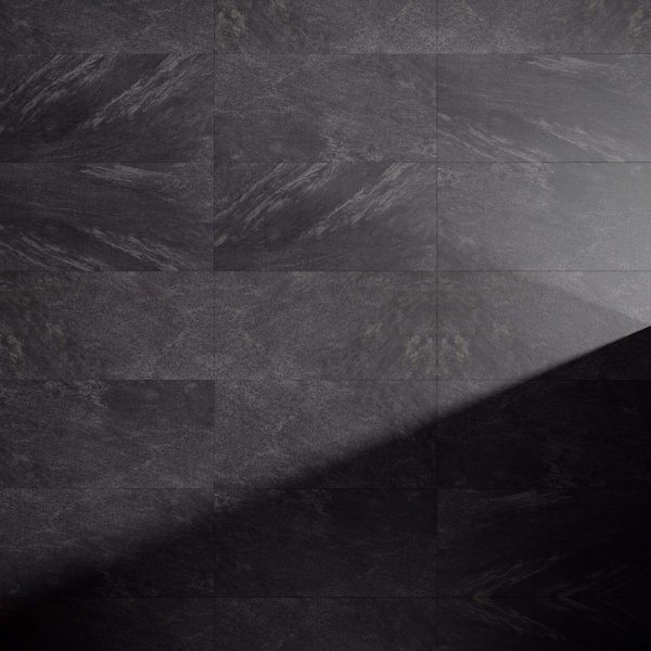 Anthracite tiles with a matte finish are lit by a diagonal beam from the lower right, highlighting their subtle natural textures and patterns, and creating a striking contrast between illuminated and shadowed areas.
