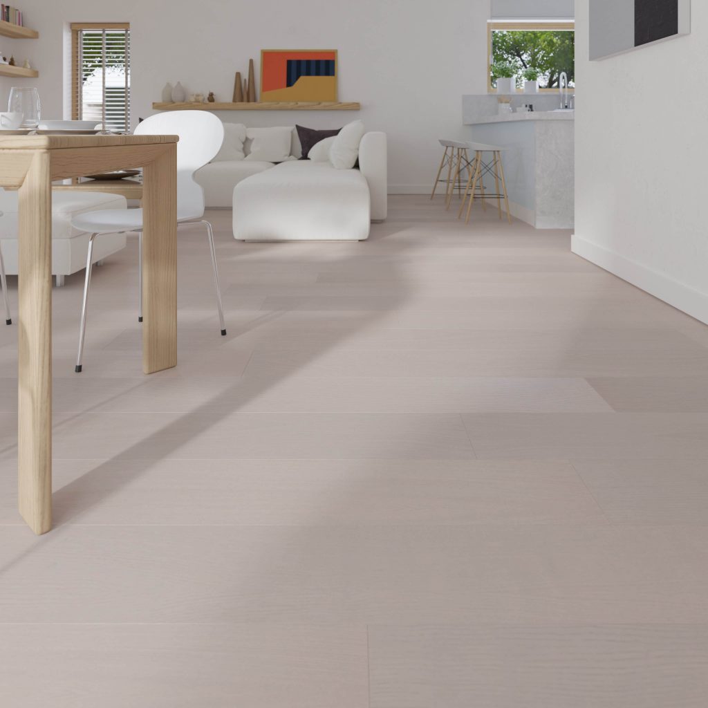 A modern, minimal living room features Braeburn Oak R&Q floors, a white sofa, wooden dining table, white chairs, and a small kitchen area. Sunlight through the windows creates a bright, airy atmosphere.