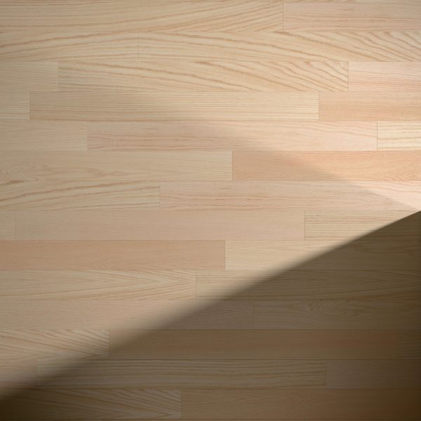Red Oak Natural* flooring features a light wood tone with a natural grain pattern. A diagonal shadow stretches from the bottom right, highlighting the contrast between sunlit and shaded sections of the floor.