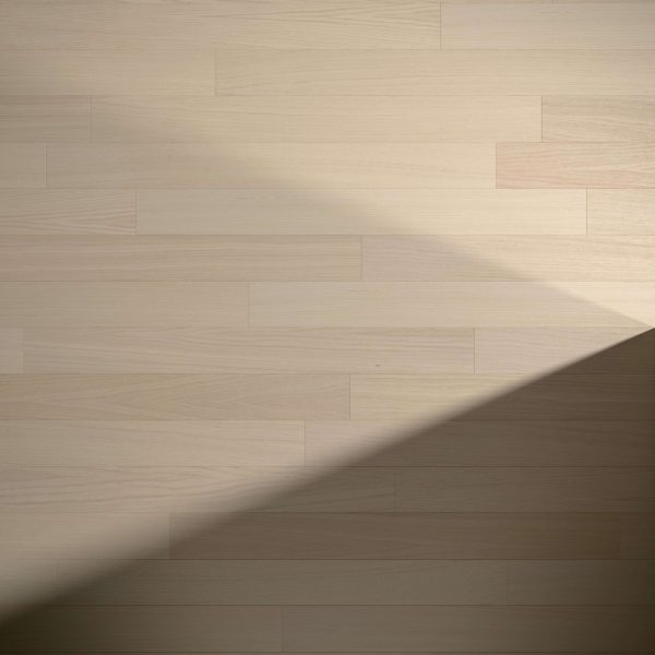 White Oak Natural* flooring features light wooden planks laid horizontally. A diagonal shadow extends from the bottom right corner toward the center, highlighting the contrast between illuminated and shaded areas on the surface.