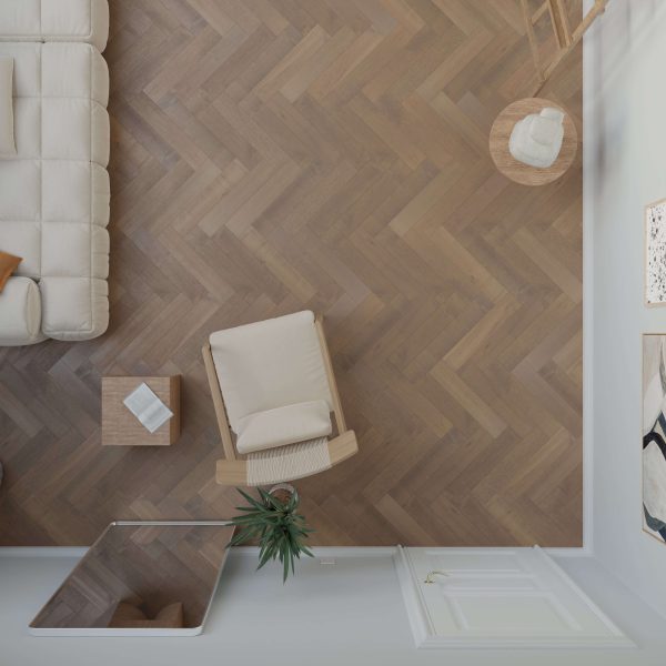 A top-down view of a modern living room features Maple Arabica* (Left) herringbone flooring, a beige armchair, side table with a book, part of a white sofa, a plant, and a closed white door.
