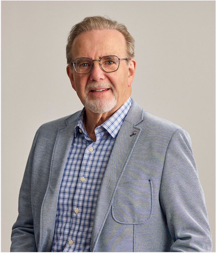 An older man with gray hair, glasses, and a trimmed beard wears a light blue blazer over a blue plaid shirt, standing against a plain background inspired by the clean look of TORLYS flooring and smiling slightly at the camera.