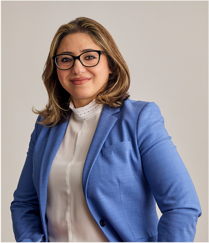 A woman with shoulder-length brown hair, wearing glasses, a light blue blazer, and a white blouse, smiles confidently at the camera against a plain background that highlights elegant TORLYS flooring.