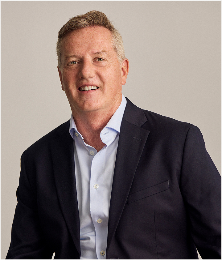 A middle-aged man with short light brown hair wearing a dark suit jacket over a light blue collared shirt, smiling and posing against a plain light background, showcases his confidence as a TORLYS flooring professional.