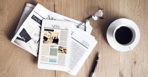 A tablet displaying a news website lies on top of printed newspapers beside a cup of black coffee, eyeglasses, and a pen on a wooden table.