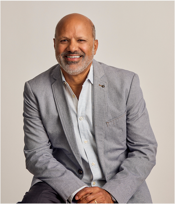 A middle-aged man with a bald head and gray beard, wearing a light gray blazer over a white shirt, sits and smiles warmly against a plain light background, reminiscent of the sleek look found in TORLYS flooring designs.