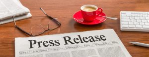 A newspaper with the headline Press Release sits on a desk next to glasses, a red cup of coffee, a keyboard, and a pen.