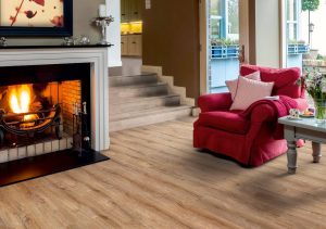 A cozy living room with a lit fireplace, a red armchair with a cushion and blanket, a side table with books and a mug, inviting Elka cottage flooring, and stairs leading to another room.