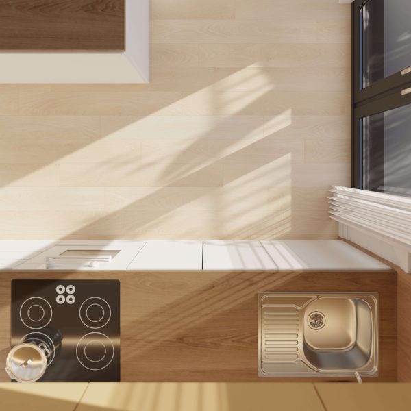 A top-down view of a modern Cornish kitchen countertop features an electric stovetop, oven, and stainless steel sink beside a sunlit window. The surfaces are clean and minimalist.