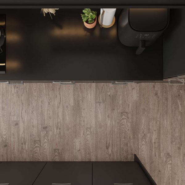 Overhead view of a modern kitchen counter with dark surfaces, a potted plant, a roll of paper towels, and the Foremast air fryer. The counter faces a wood-patterned floor.