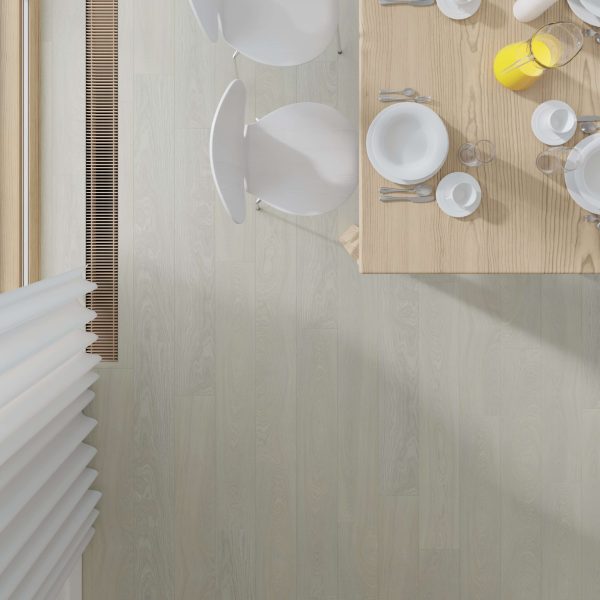 A top-down view of a Somerville Oak dining table set for breakfast with plates, cups, cutlery, a glass jug of orange juice, and three white chairs on a pale wood floor near a window with vertical blinds.