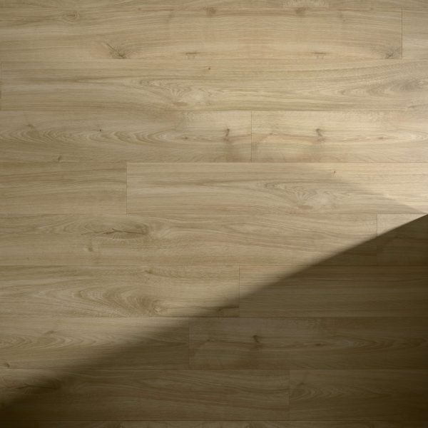 Juventas Oak light brown wooden floor features visible grain patterns. A diagonal shadow crosses the lower right area, adding contrast between illuminated and shaded sections.