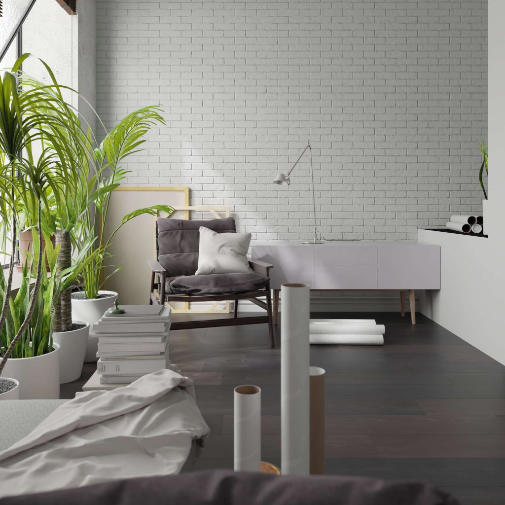 A modern, bright room with white brick walls, Beaumont Oak floors, potted green plants, a gray cushioned chair with pillows, a white desk, and various rolled papers and art supplies scattered around.