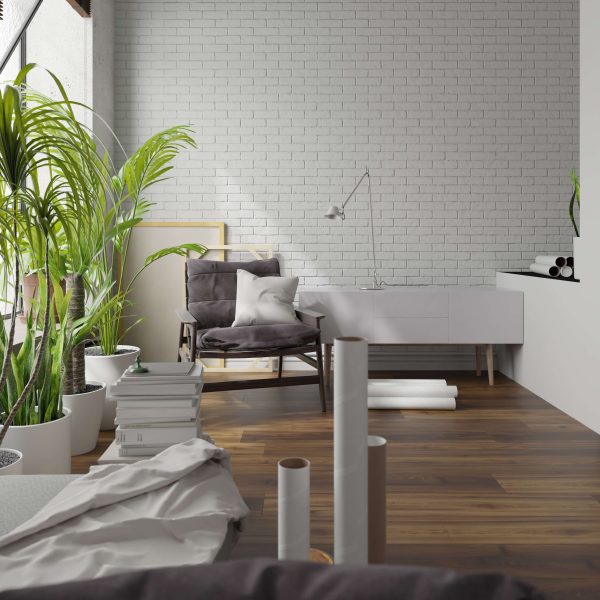 A modern, bright room with a white brick wall and Walcourt Hickory wooden floor, potted plants, a cushioned gray chair, white sideboard, and books—creating a cozy, artistic workspace.