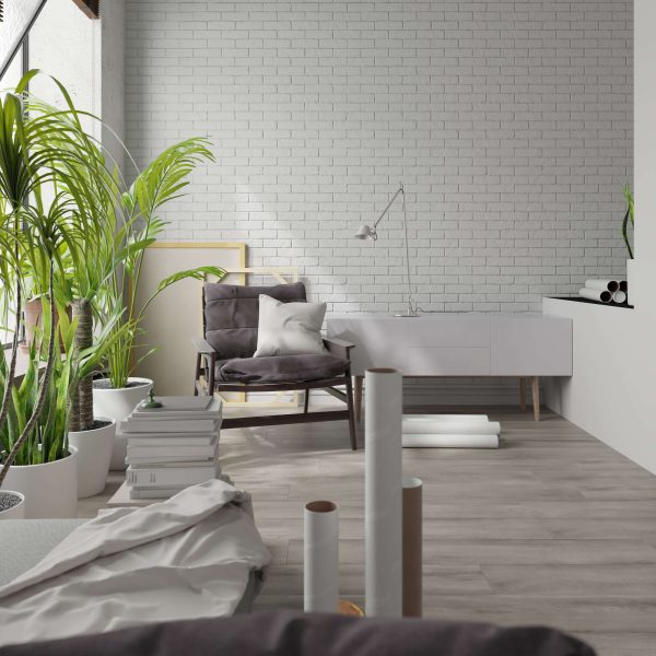 A modern, bright room features Rochefort Oak floors, white brick walls, potted plants, a gray cushioned chair with pillows, a white sideboard, and rolled-up papers scattered throughout the space.