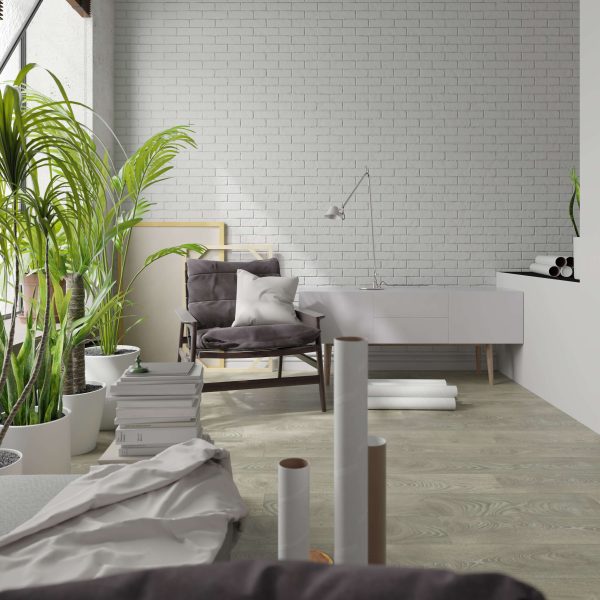 A modern, bright room features a Landen Oak gray armchair, white desk, potted plants, stacked books, rolled papers, and a white brick wall. Natural light from the left creates a cozy and fresh atmosphere.