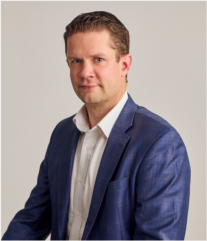 A man with short brown hair wearing a blue plaid suit jacket and white dress shirt poses against a plain, light-colored background, reminiscent of the clean lines and modern style of TORLYS flooring. He faces slightly right, looking at the camera with a neutral expression.