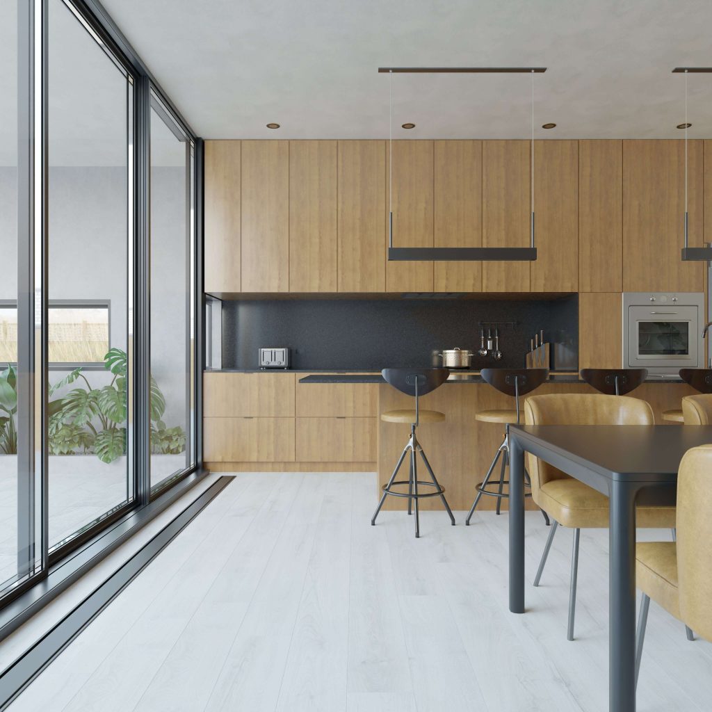 The Elkington kitchen features wood cabinets, black countertops, a built-in oven, bar stools, a dining table, and large glass doors that bring in abundant natural light. Green plants can be seen outside for a fresh view.