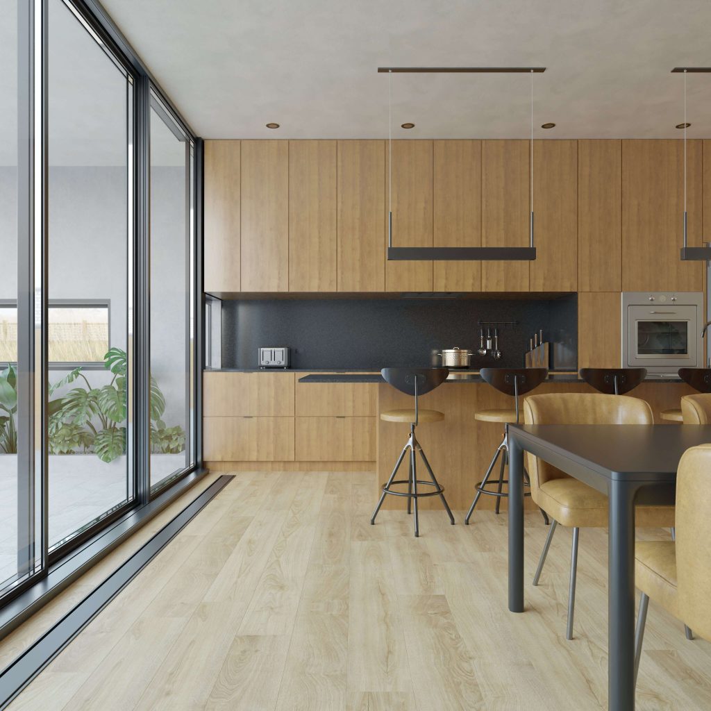 The Brightstone kitchen features light wood cabinets and floors, black countertops, a built-in oven, bar stools, a dining table with brown chairs, and large glass doors filling the space with natural light. Lush green plants are visible outside.