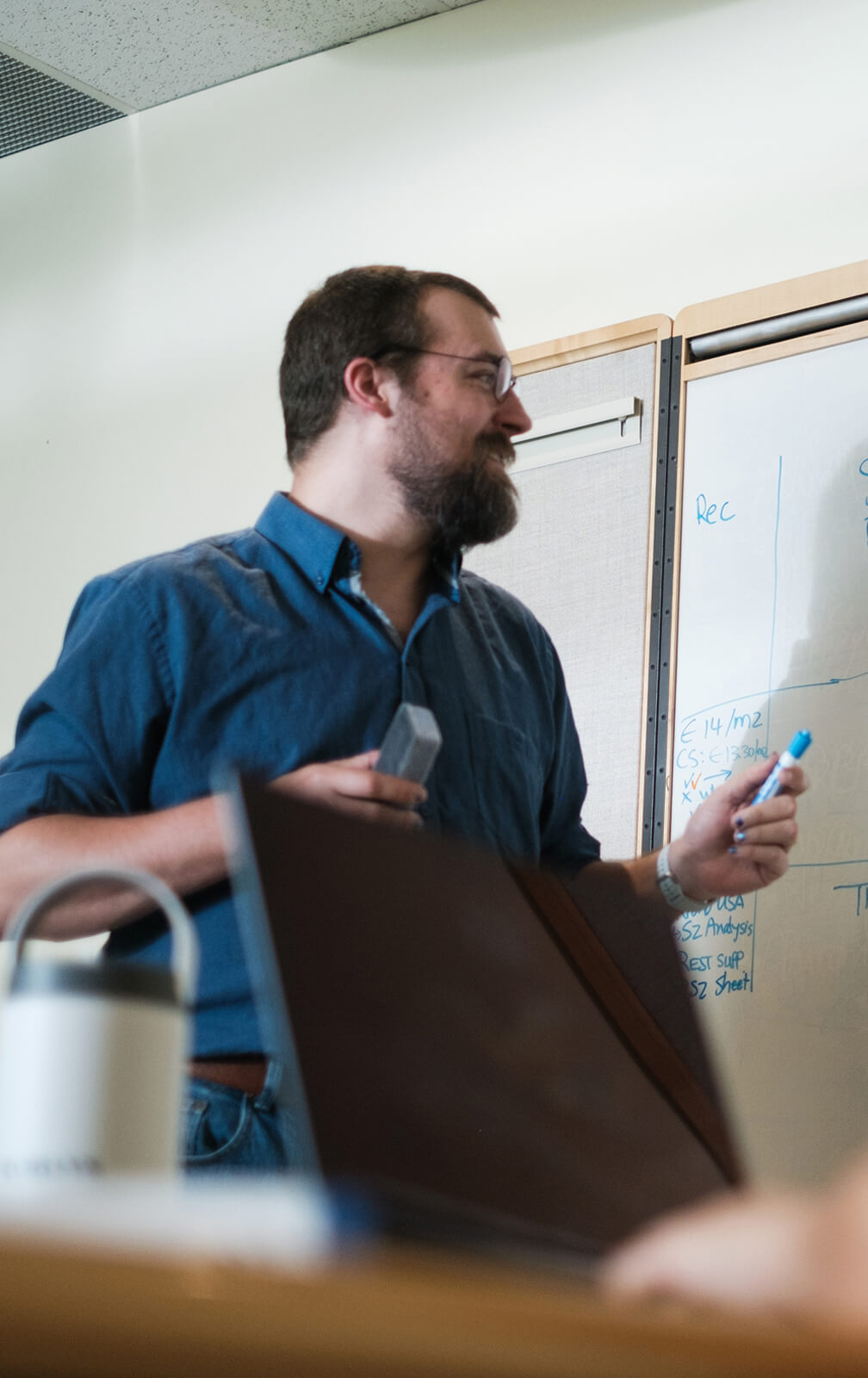 A man with glasses and a beard stands by a whiteboard holding markers, engaged in discussion. A laptop, coffee mug, and TORLYS flooring samples are visible in the foreground. The whiteboard has handwritten notes.