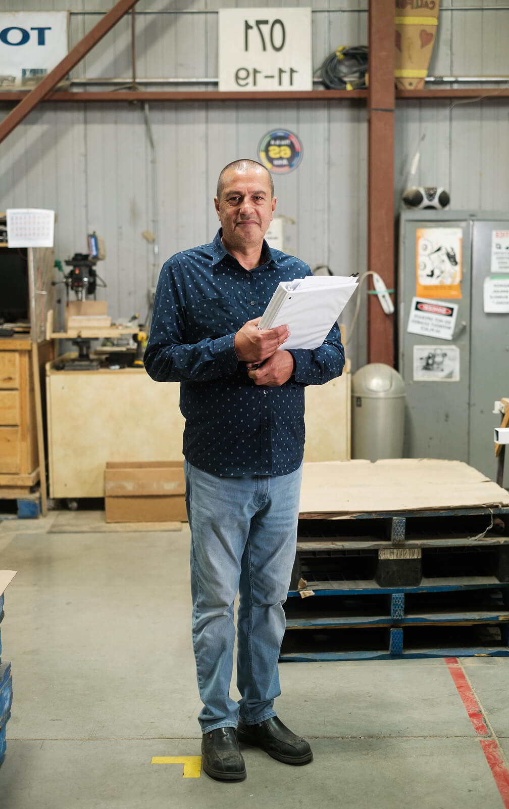 A man in a blue shirt and jeans stands indoors holding papers, with industrial shelves, equipment, and signs behind him.