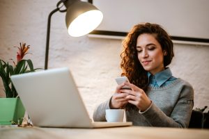 A woman with curly hair sits at a desk using her smartphone, with an open laptop in front of her, a coffee cup, a plant, and a lit desk lamp nearby, adding to her relaxed demeanour.