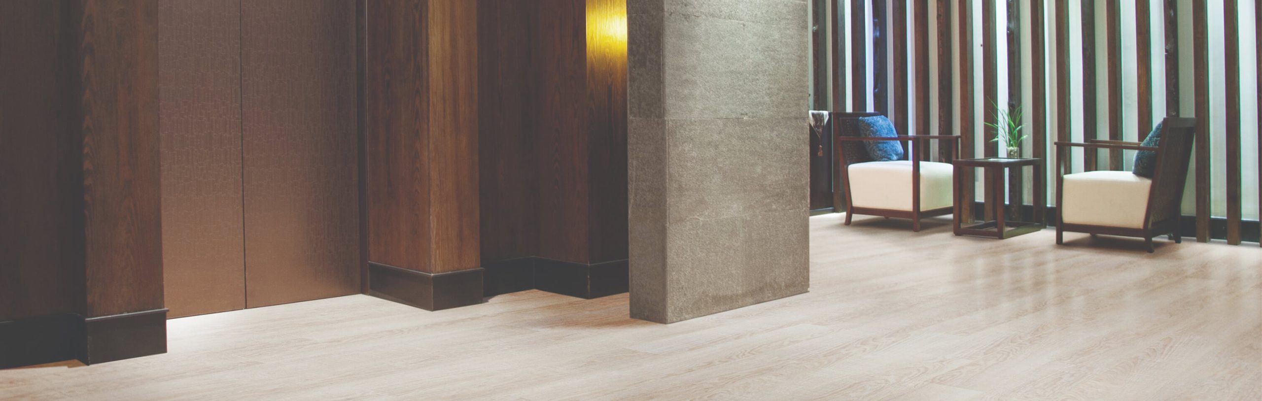 Modern lobby area managed by Floors Property Management, featuring light wood flooring, dark wooden wall panels, a stone pillar, and a cozy seating area with armchairs and a small table by vertical slats bathed in natural light.