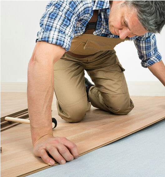 A person in a plaid shirt and khaki overalls kneels on the floor, installing wooden flooring panels, with tools nearby.