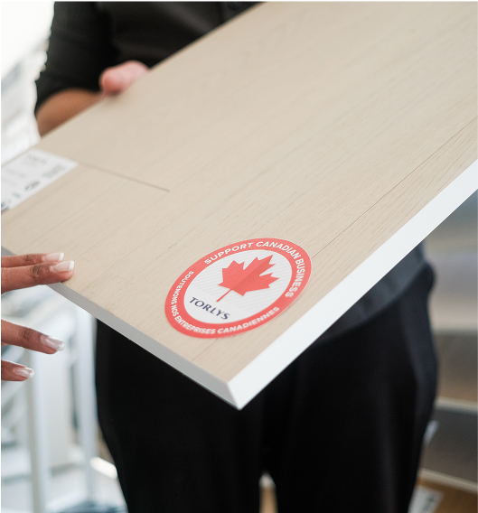 A person holds a light wood panel with a red and white Support Canadian Business sticker featuring a maple leaf and the name Torlys. Another hand reaches toward the panel.