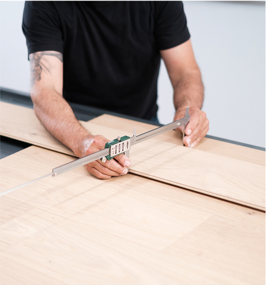 A person in a black shirt uses a digital caliper to measure the width of a light wooden board on a work surface.