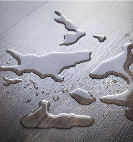 Water droplets and small puddles scattered on a dark wooden surface, showing reflections and the texture of the wood grain.