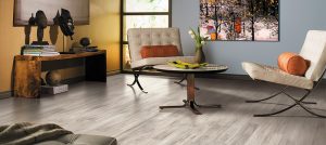A modern living room with light gray wood flooring, two white cushioned chairs with orange pillows, a round coffee table, a wooden console with decor, and a colorful painting on the wall.