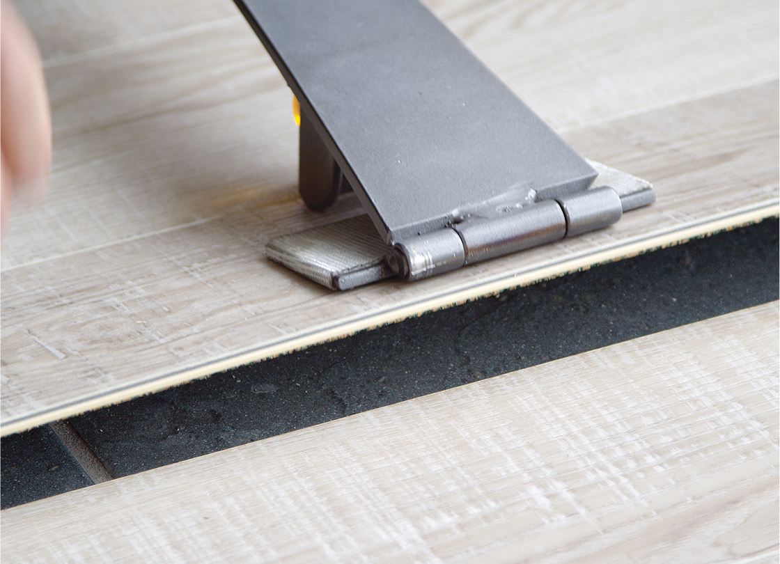 A close-up of a Smart Floors Accessories tool being used to install or adjust a piece of light-colored laminate flooring over black underlayment. The tool presses the flooring plank firmly in place for a seamless fit.
