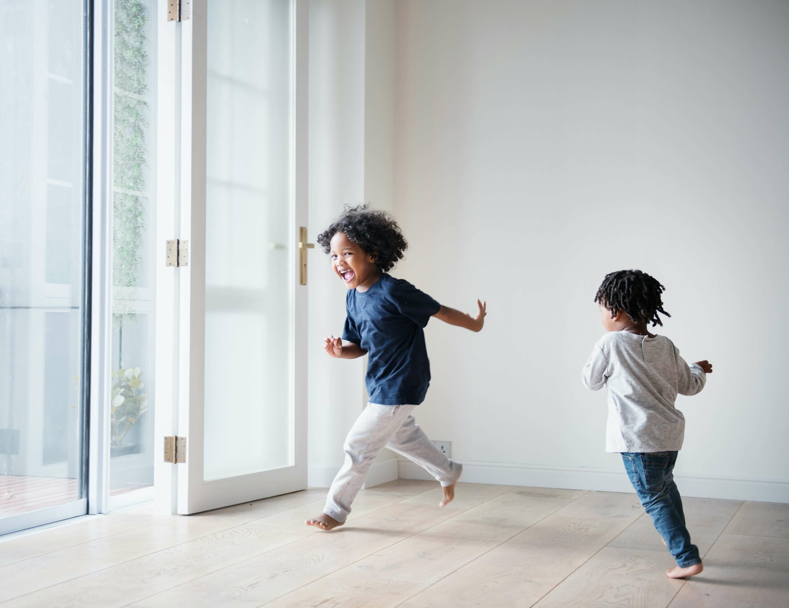 Two young children joyfully run barefoot across a sustainable wooden floor in a bright room with large glass doors, letting in natural light.