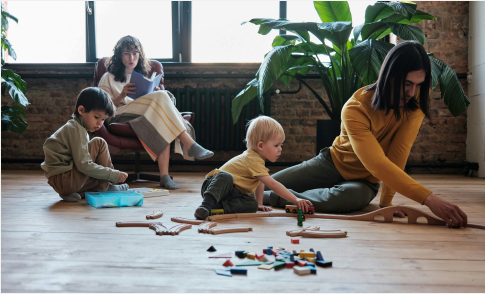 Two children play with wooden toys on the floor while an adult helps them. Another adult sits in the background on a chair, reading a book. Large green plants and natural light fill the cozy room.